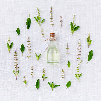 Glass bottle of essential oil surrounded by fresh herbs on white linen
