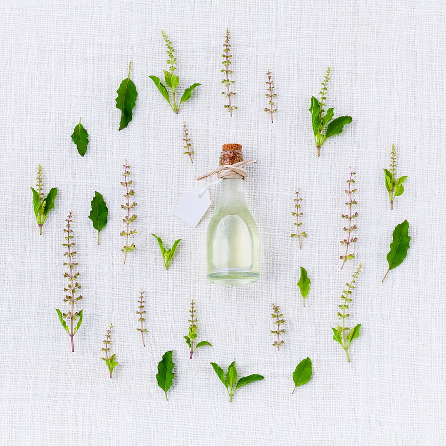 Glass bottle of essential oil surrounded by fresh herbs on white linen