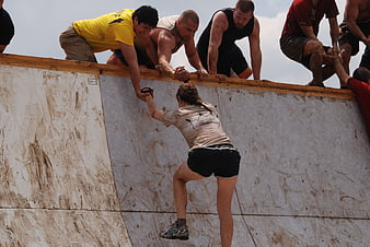 Woman climbing muddy wall with team help at obstacle course