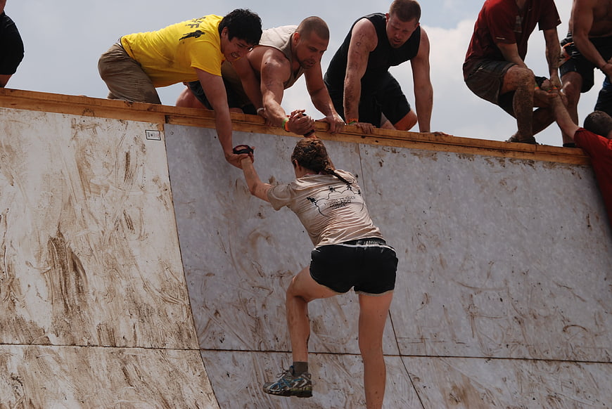 Woman climbing muddy wall with team help at obstacle course