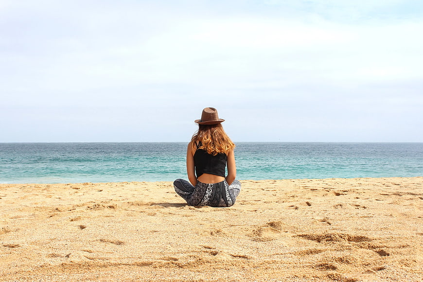 Woman in Hat Sitting on Sandy Beach Facing Ocean