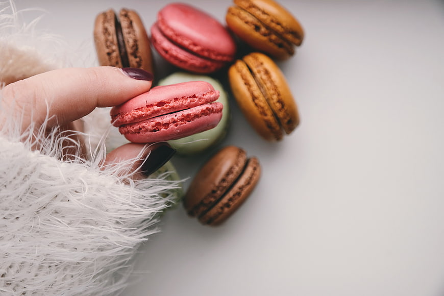 Hand holding pink macaron with assorted pastries on white background