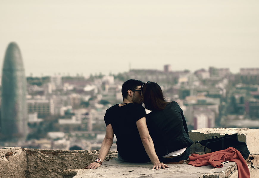 Romantic Couple Kissing On Stone Wall With Blurred Barcelona Cityscape Background