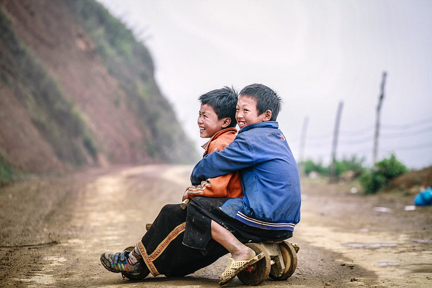 Two smiling children riding a wooden cart on a dirt road