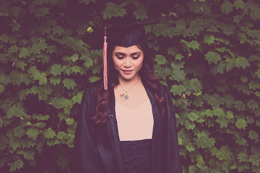 Female graduate in black gown and cap with pink tassel against green ivy background