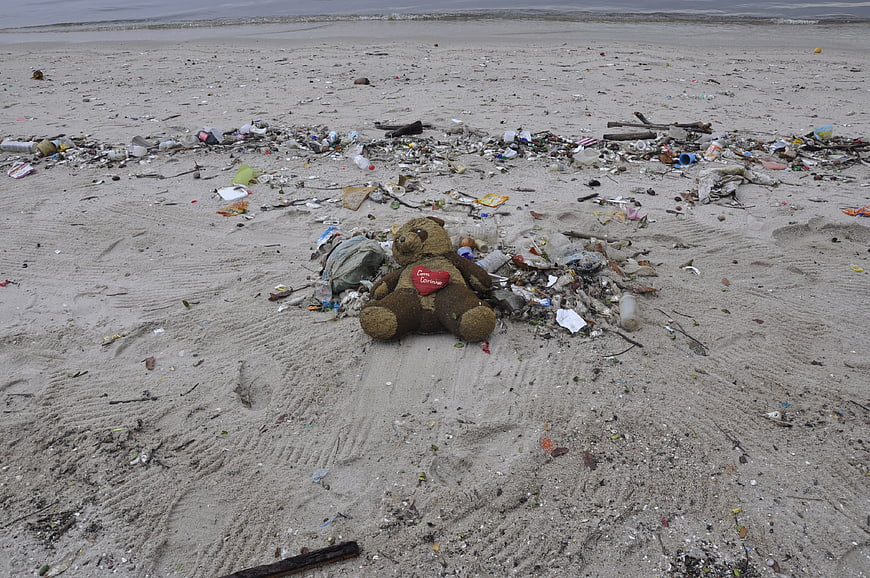 Abandoned Teddy Bear Surrounded By Plastic Trash On Polluted Beach