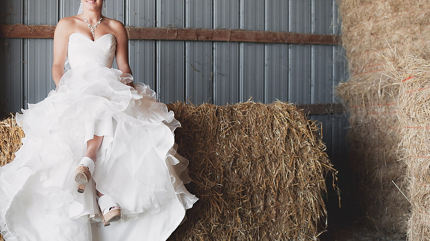 Bride in White Gown Sitting on Hay Bale in Barn