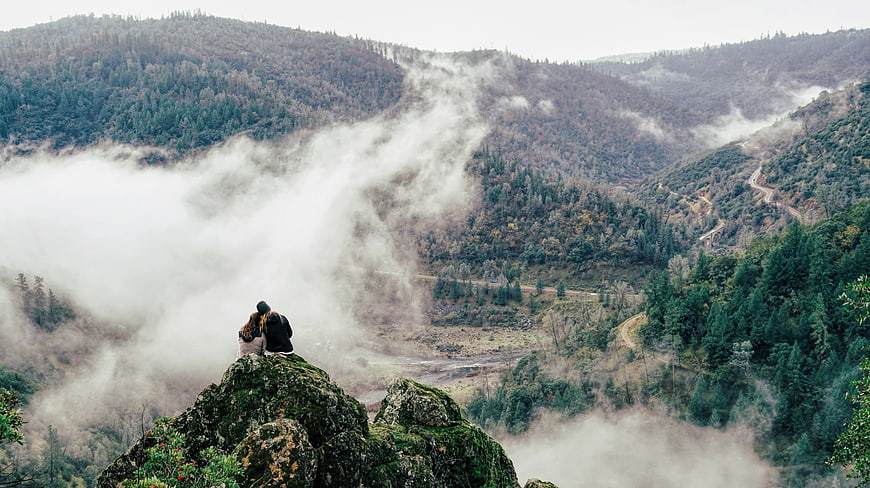 Couple sitting on rocky cliff overlooking misty mountain valley