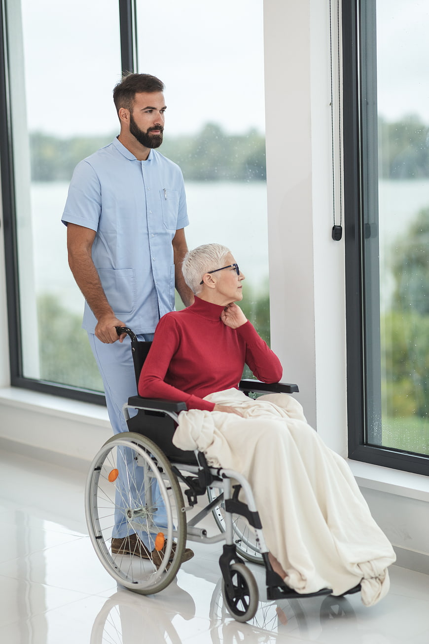 Male nurse pushing elderly woman in wheelchair by window