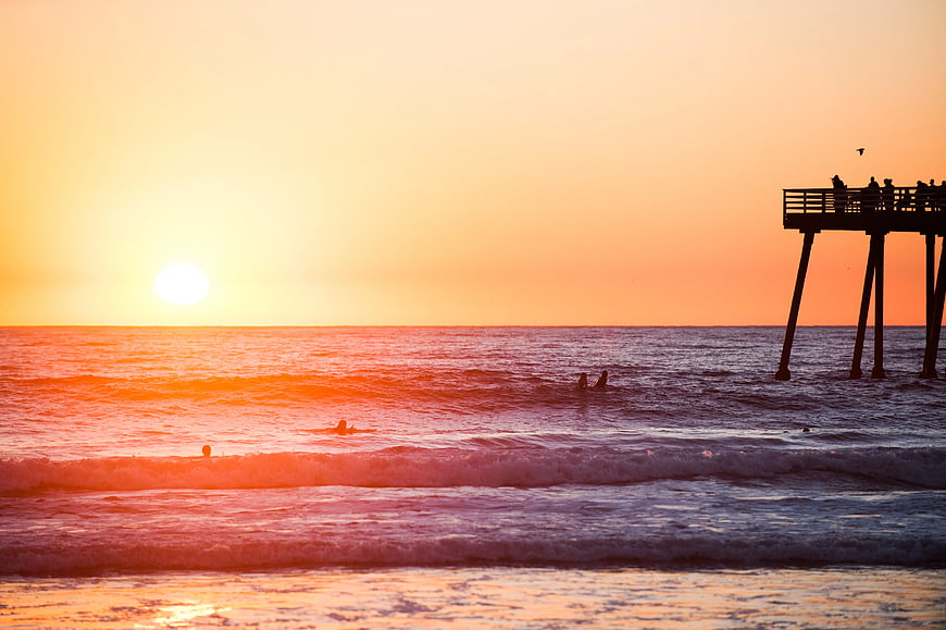 Sunset over ocean with pier silhouette and surfers