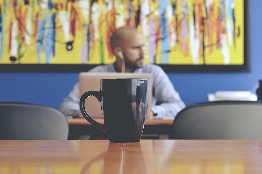 Black coffee mug on table with blurred man working on laptop