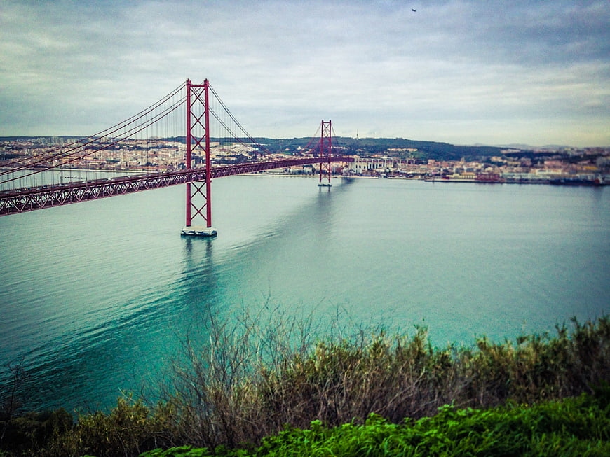 Red suspension bridge spanning river with cityscape background