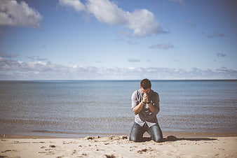 Man Kneeling in Prayer on Sandy Beach by Ocean