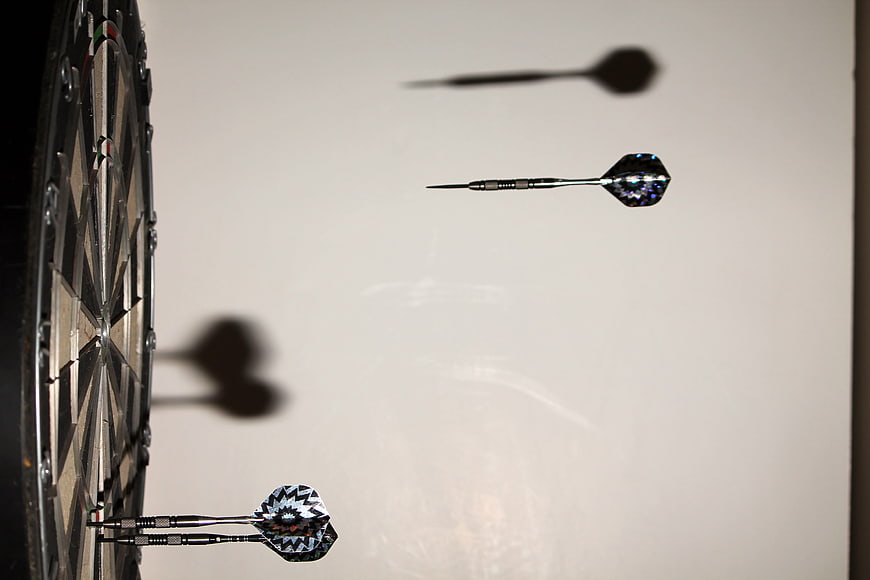 Darts in mid-air with shadows on a wall near a dartboard