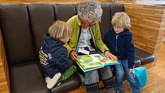 Grandmother Reading Picture Book To Two Young Boys In Library