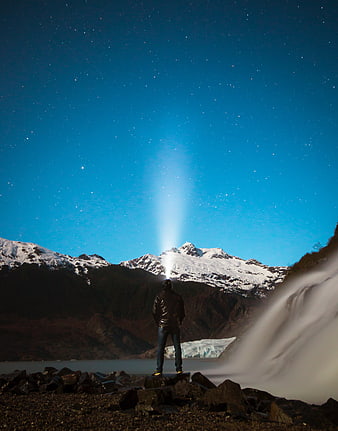 Man with flashlight beam under starry night sky near waterfall