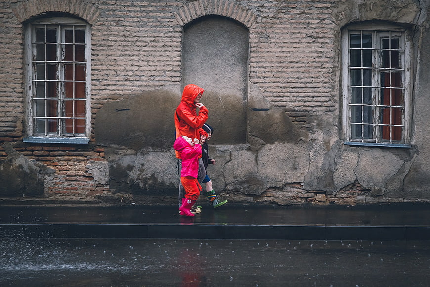 Family in Raincoats Walking Past Old Brick Wall
