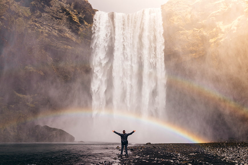 Man with arms raised facing massive waterfall and rainbow