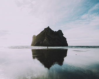 Solitary figure standing on wet beach with rock formation reflection