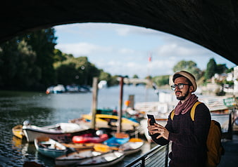 Man with hat and backpack using smartphone by river boats