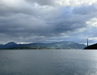 Dramatic suspension bridge over water with mountains and cloudy sky
