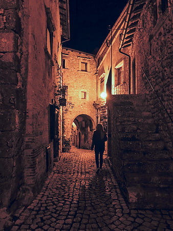 Woman Walking Down Narrow Cobblestone Alley at Night