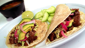 Two beef tacos with avocado, pickled onions, and cucumber on a white plate
