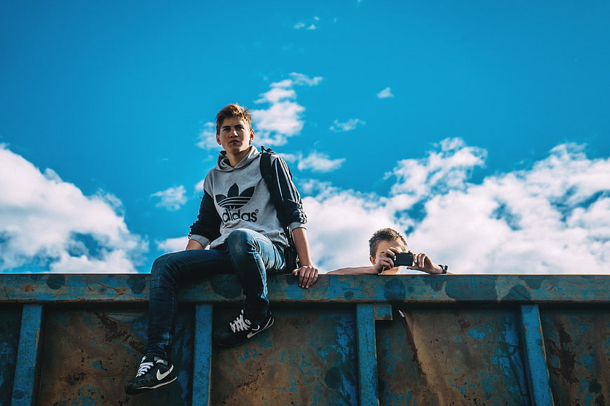 Two boys sitting on a rusty blue container against a sky