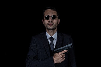 Man in Suit and Sunglasses Holding Pistol Against Black Background