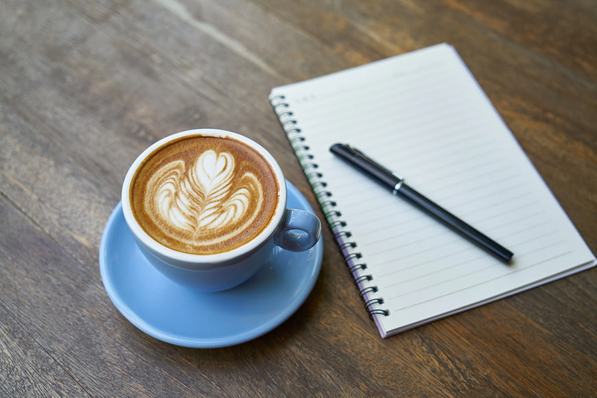 Latte with heart art beside notebook and pen on wood