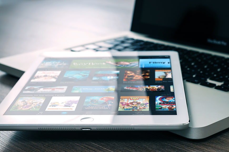 White tablet displaying movie apps on wooden desk with laptop