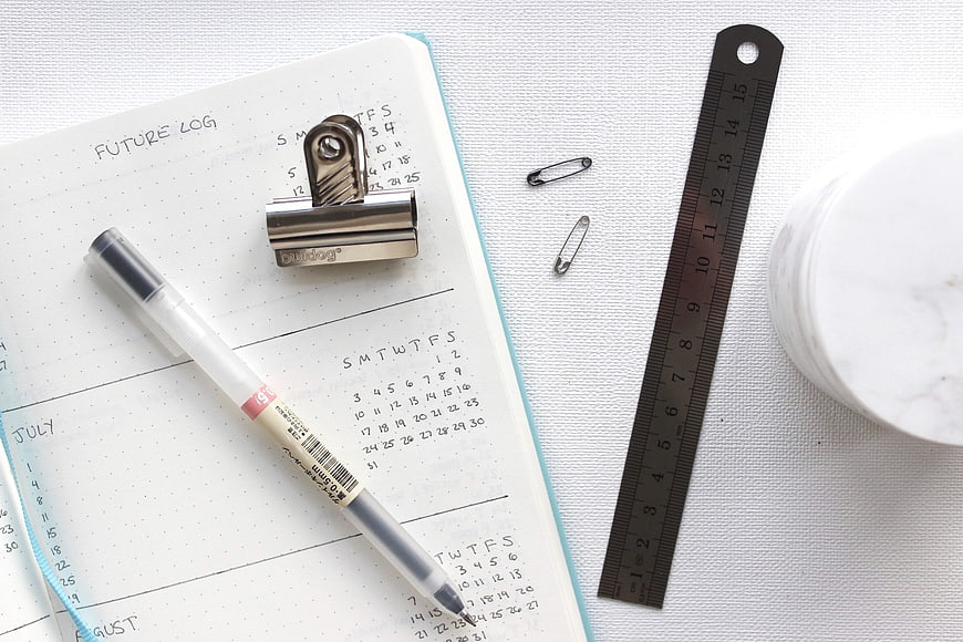 Flat lay of bullet journal, pen, ruler, and safety pins on white background