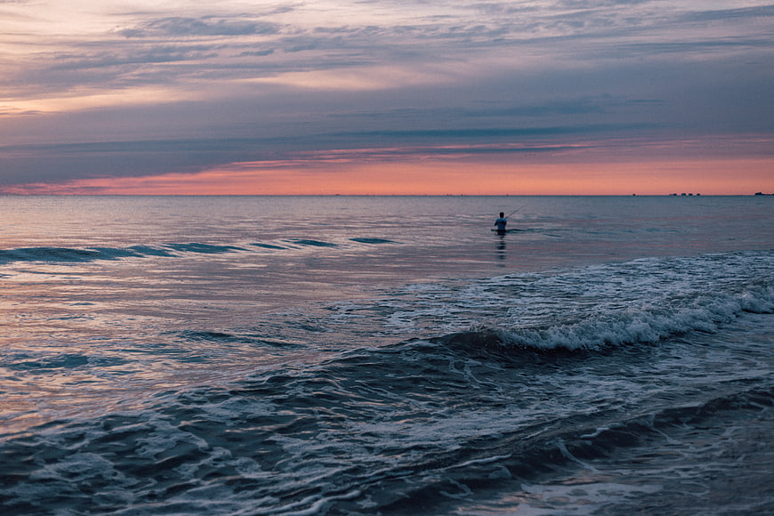 Lone fisherman in ocean waves during pink sunset