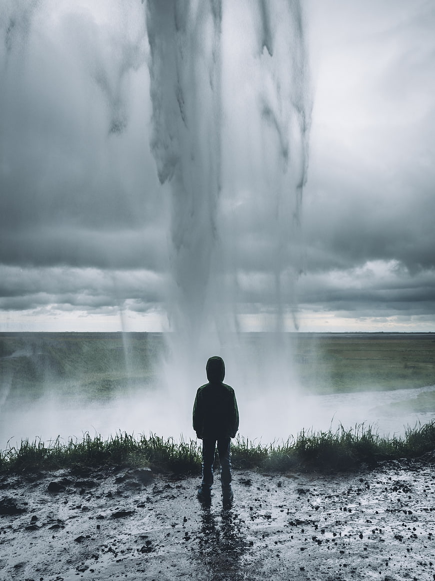 Lone figure standing before a massive waterfall in misty landscape