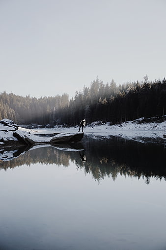 Lone figure standing on snowy rock by calm winter lake