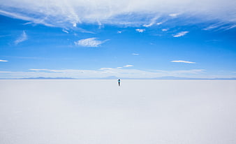 Lone figure walking on vast white salt flat under blue sky