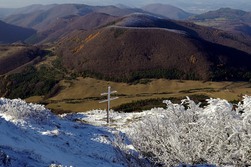 Snow-covered wooden cross on mountain ridge overlooking autumn valley