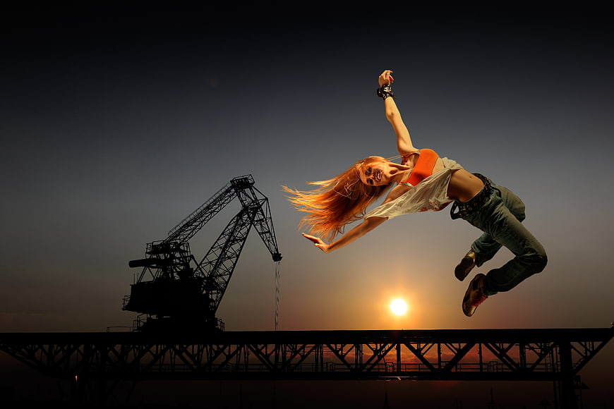Woman performing acrobatic jump against sunset and industrial crane