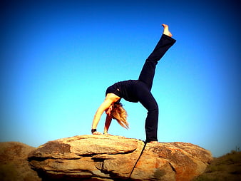 Woman performing advanced yoga backbend on a large rock against a blue sky