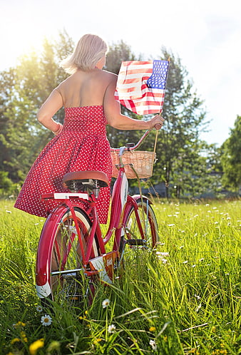 Senior Woman On Red Bicycle Holding American Flag In Summer Meadow