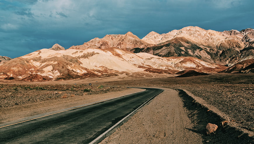 Winding desert road leading to colorful mountains under dramatic sky