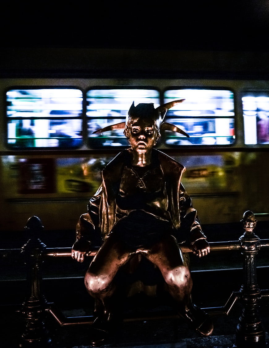 Bronze Joker Statue Sitting on Bench with Blurred Tram Background