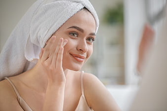 Woman with towel on head applying skincare in bathroom mirror