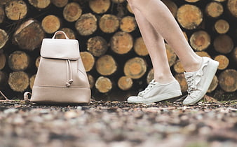 Woman's legs in white sneakers near beige backpack and wood logs