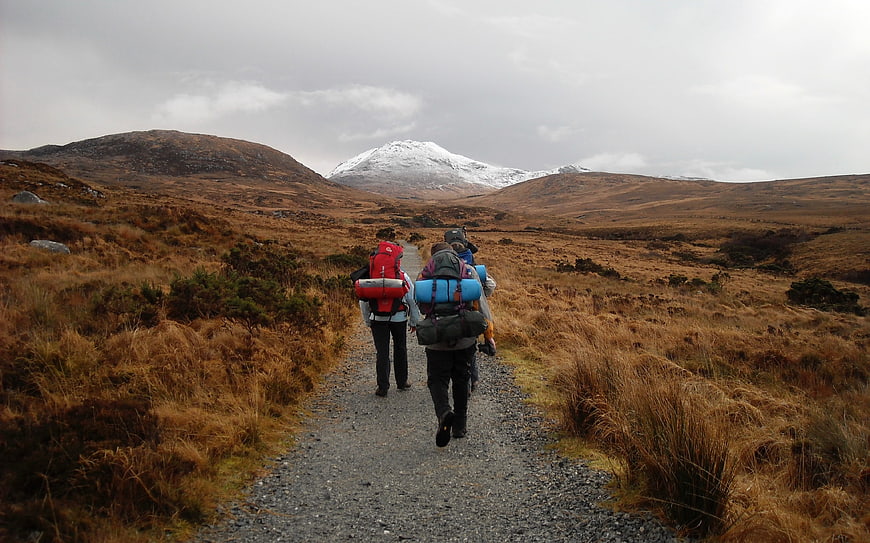 Hikers with backpacks walking on a gravel path toward a snow-capped mountain