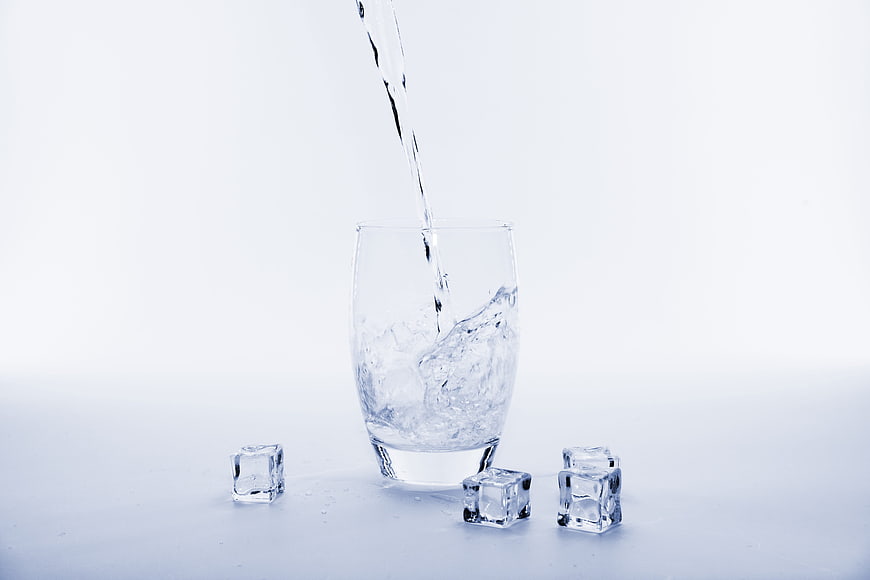 Water Pouring Into Glass With Ice Cubes On White Background