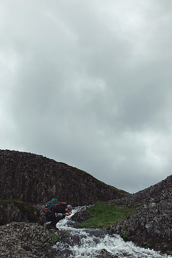 Hiker with backpack crouching by mountain stream under cloudy sky