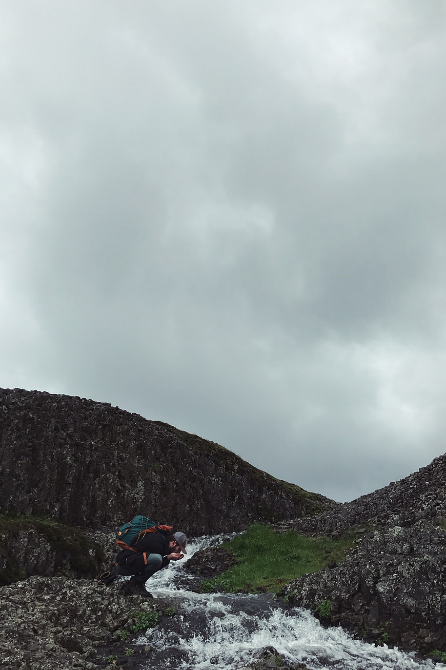 Hiker with backpack crouching by mountain stream under cloudy sky