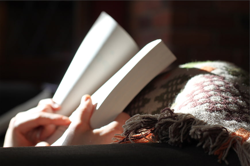 Close-up of hands reading a book under a cozy blanket