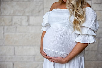 Pregnant woman in white dress holding belly against stone wall